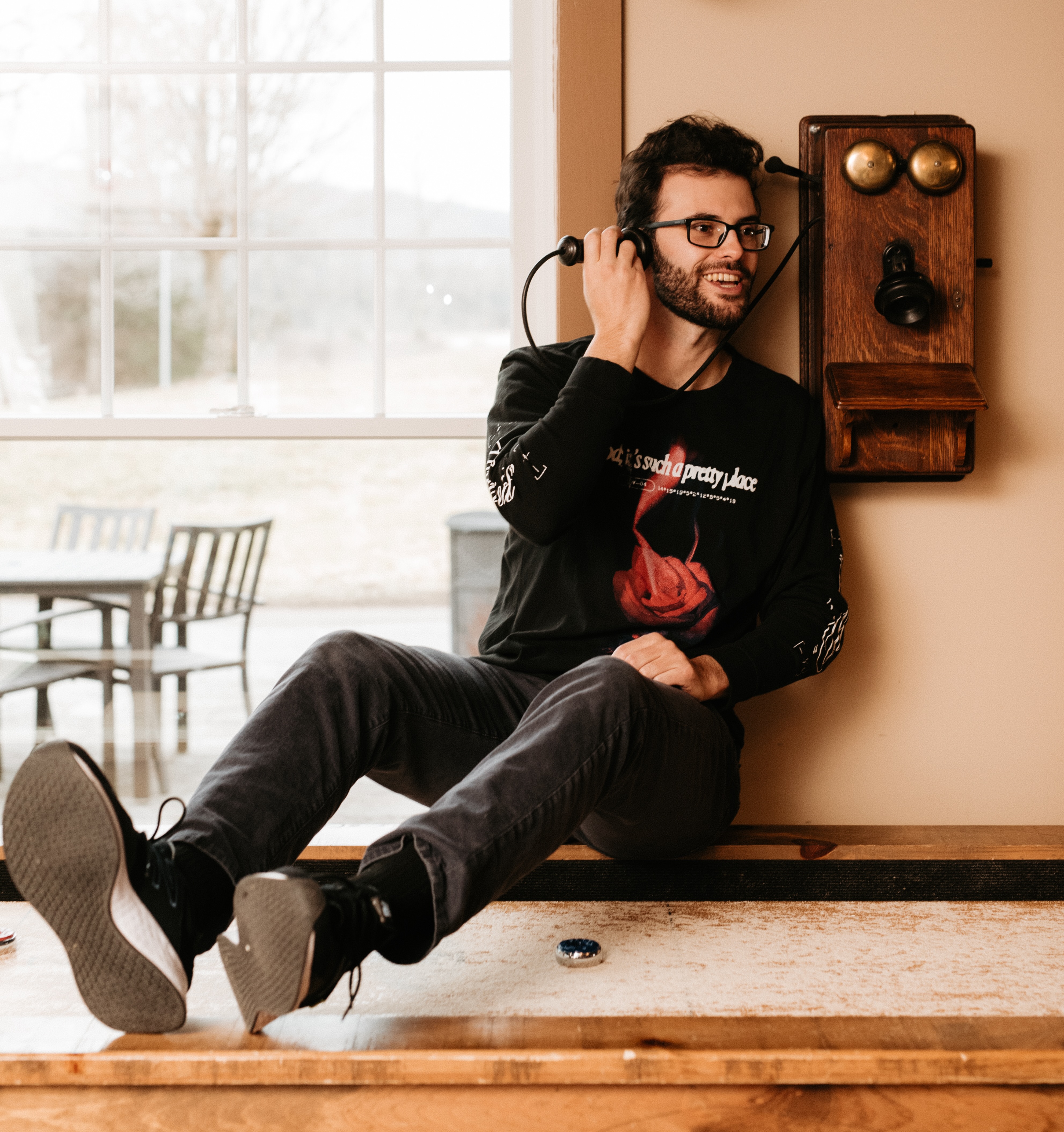 Matt sits on top of a shuffleboard table, jokingly holding a not-plugged-in, old-timey telephone up to his ear